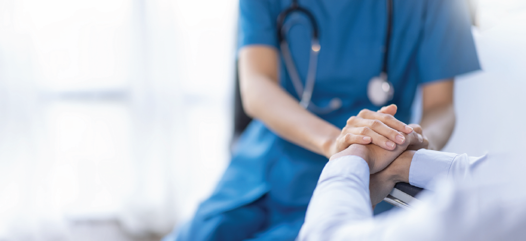 A nurse in a blue uniform with a stethoscope holding a senior patient’s hand.