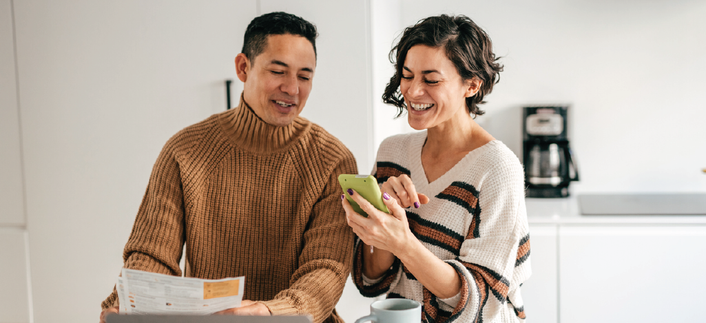 Text and email bill notifications - Couple at home paying bills online using a smartphone and laptop.