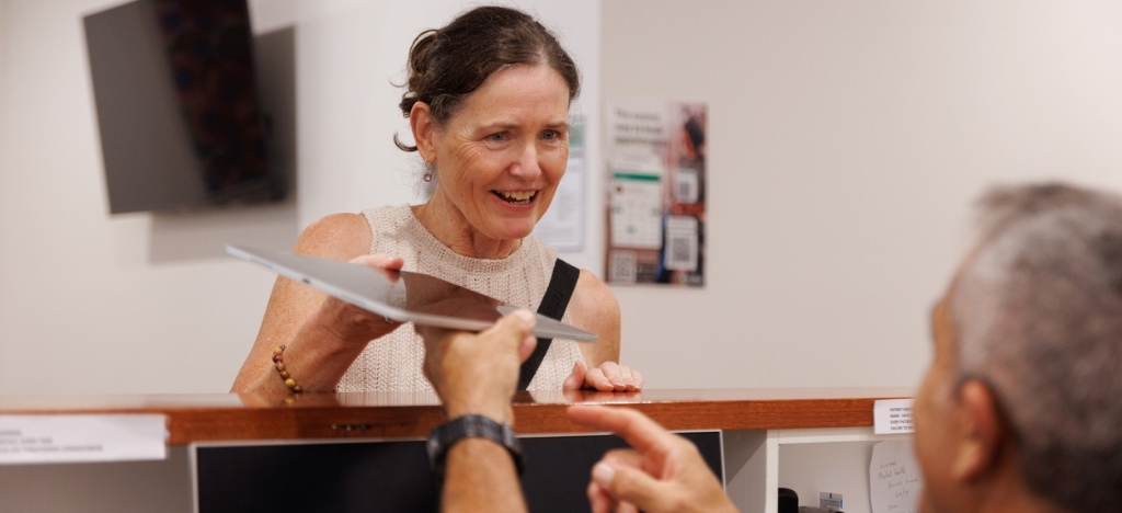 Female patient hands digital tablet to male front desk medical staff.
