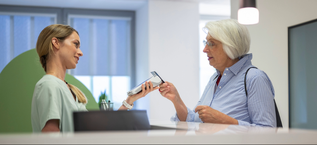 Front-desk staff hold a payment terminal for an elderly lady to tap their card for payment.