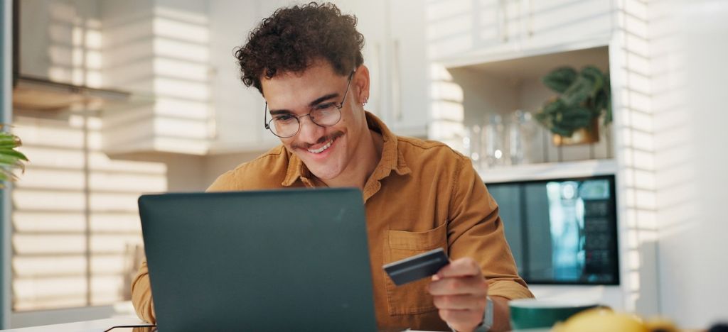 Man smiles as he holds a card while paying on a laptop at home.