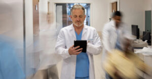 Managing patient surge - A male doctor holding a tablet device while standing in a busy hospital hallway.