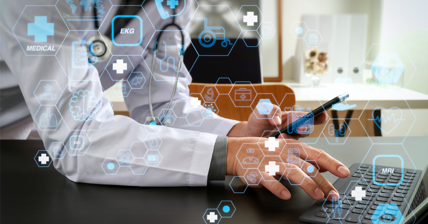 Upgrading healthcare technology stack - A male doctor using his phone and computer to communicate patient treatment procedures.