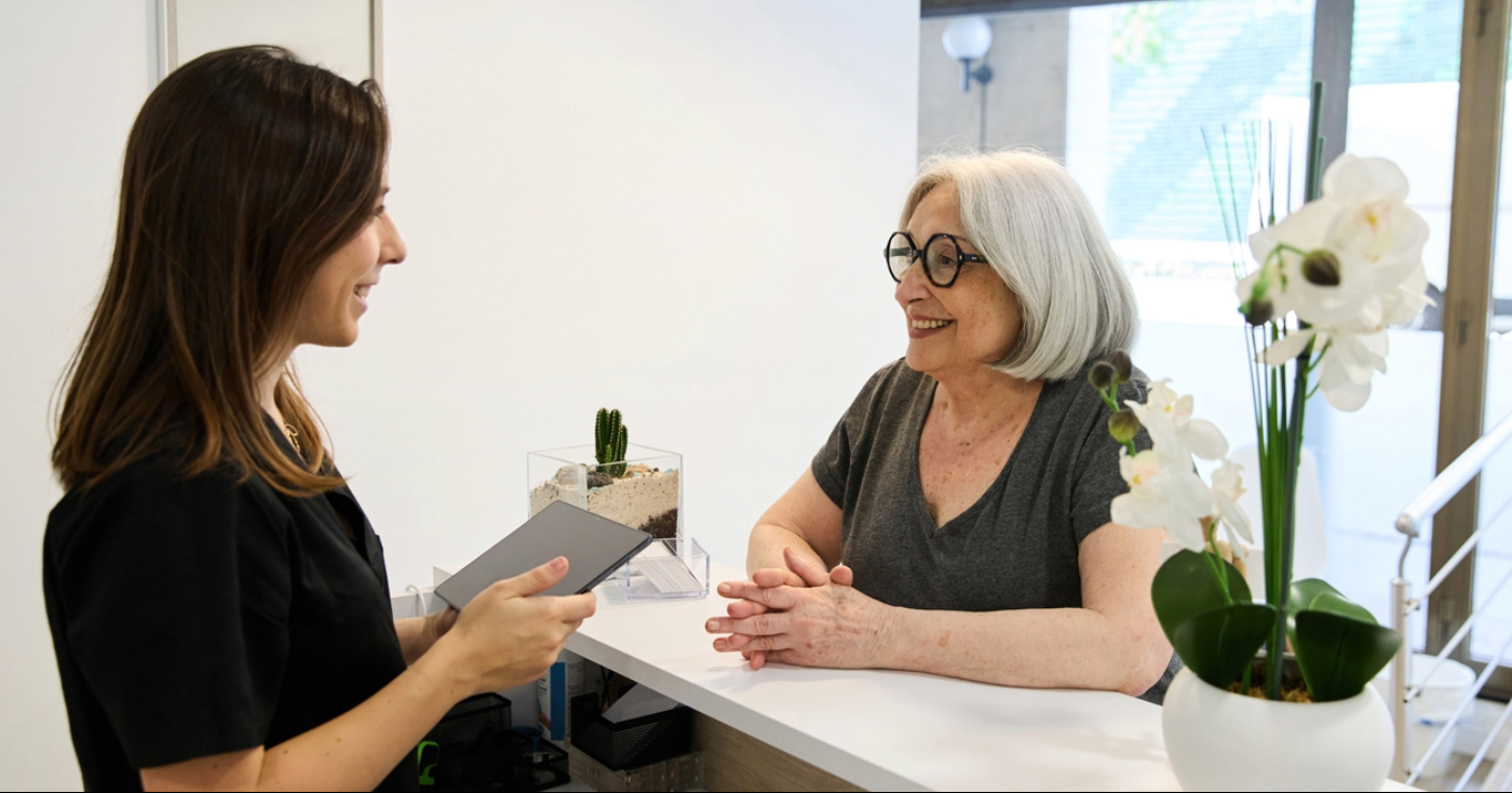 Female medical receptionist smiling while assisting a patient at the front desk.